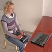 Frau mit Stenografie-Tastatur zwischen den Knien sitzend vor Tisch mit Laptop., Grossansicht öffnen