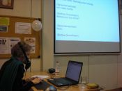 Frau mit Maske über Mund sitzt vor Laptop und schreibt. Im Hintergrund eine Beamerleinwand mit Schrift., Grossansicht öffnen