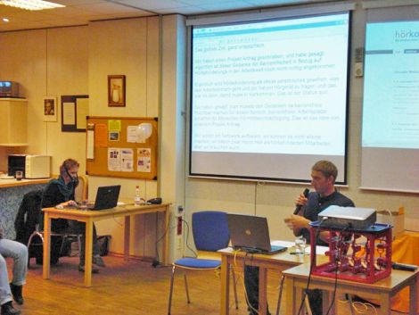 Veranstaltungssituation mit einem Sprecher, der Mikrofon in der Hand hält. Dahinter eine Großbildleinwand mit Schrift, eine Schriftsprachdolmetschherin befindet sich im Hintergrund.